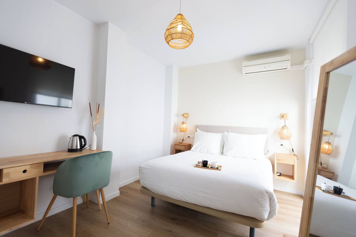 A modern hotel room with a wooden desk, a green chair, a large bed with white bedding, bedside lamps, and a flat-screen TV mounted on the wall.