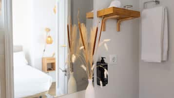 Un baño con un lavabo blanco, un espejo, un jarrón blanco con plantas secas, una estantería de madera con artículos de aseo, y una toalla blanca colgada en la pared.