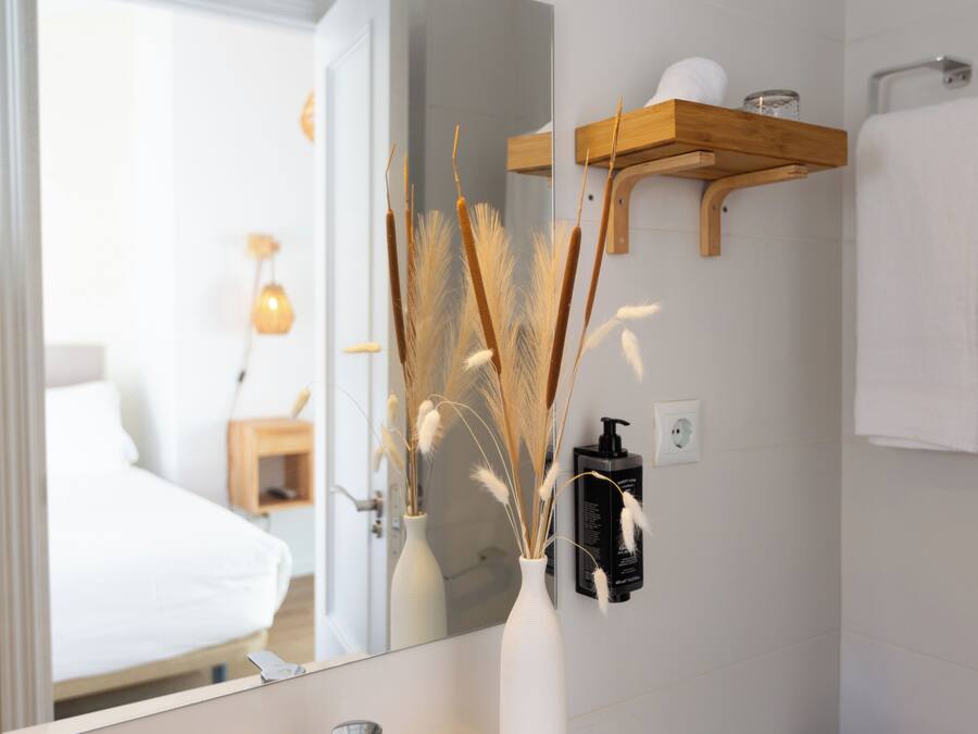 Un baño con un lavabo blanco, un espejo, un jarrón blanco con plantas secas, una estantería de madera con artículos de aseo, y una toalla blanca colgada en la pared.