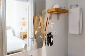 A bathroom with a white sink, a mirror, a white vase with dried plants, a wooden shelf with toiletries, and a white towel hanging on the wall. - Hostal Boutique La Malagueña Only Adults by Croma (Estepona)