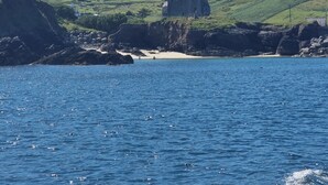Un paysage côtier avec une construction en pierre sur une colline, une plage de sable et une étendue d’eau.