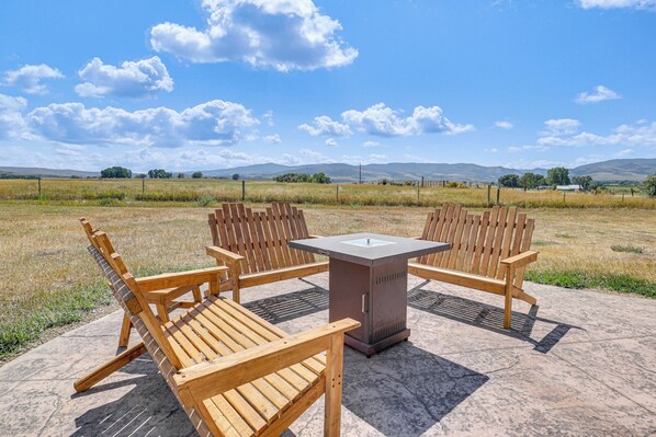 An outdoor seating area with wooden chairs and a table, set on a concrete patio with a view of a field and distant hills. - Mountain-View Log Cabin in Wyoming Wilderness (Encampment)