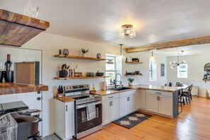 Una cocina moderna con armarios blancos, encimeras de madera y una isla central. Incluye electrodomésticos integrados, como una estufa y un horno, además de un amplio ventanal que proporciona luz natural.