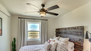 A bedroom with a bed, a ceiling fan, a window with curtains, a cactus plant, and a rustic headboard.