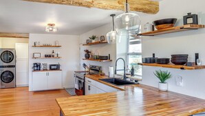 A modern kitchen with wooden countertops, a built-in oven, and a large island with a sink. There are hanging pendant lights and shelves above the counter.