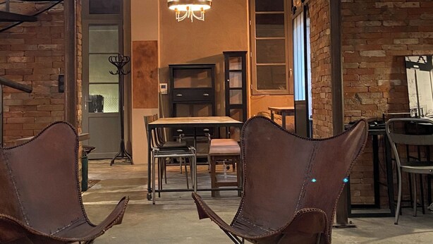 A spacious living area with a high ceiling, exposed brick walls, and a mezzanine level. There are two leather chairs and a dining table in the foreground. - Grand Loft Dartiste en Avec Équipement Professionnel Audio et Vidéo (Angoulême)