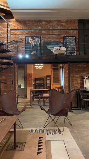Un vaste espace de vie doté d’un haut plafond, de murs en briques apparentes et d’une mezzanine. On y trouve deux fauteuils en cuir et une table à manger au premier plan.