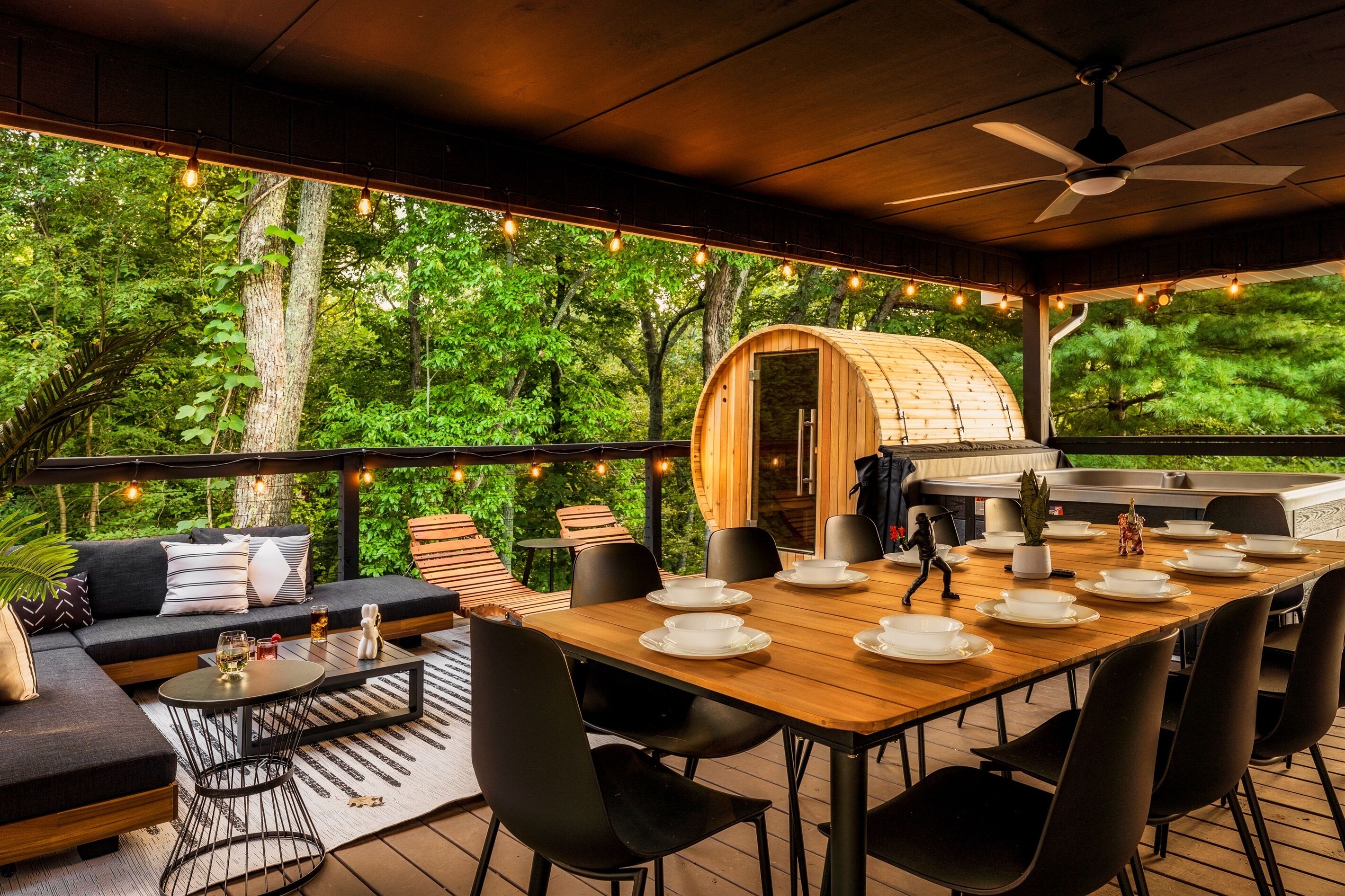 Un comedor al aire libre con una mesa de madera preparada para cuatro personas, rodeada de sillas negras, y una zona de estar acogedora con un sofá y cojines.