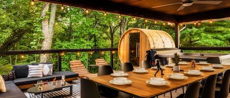 Un comedor al aire libre con una mesa de madera preparada para cuatro personas, rodeada de sillas negras, y una zona de estar acogedora con un sofá y cojines.