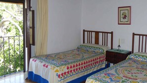 A room with two beds, a balcony with a view of greenery, and a framed picture on the wall.