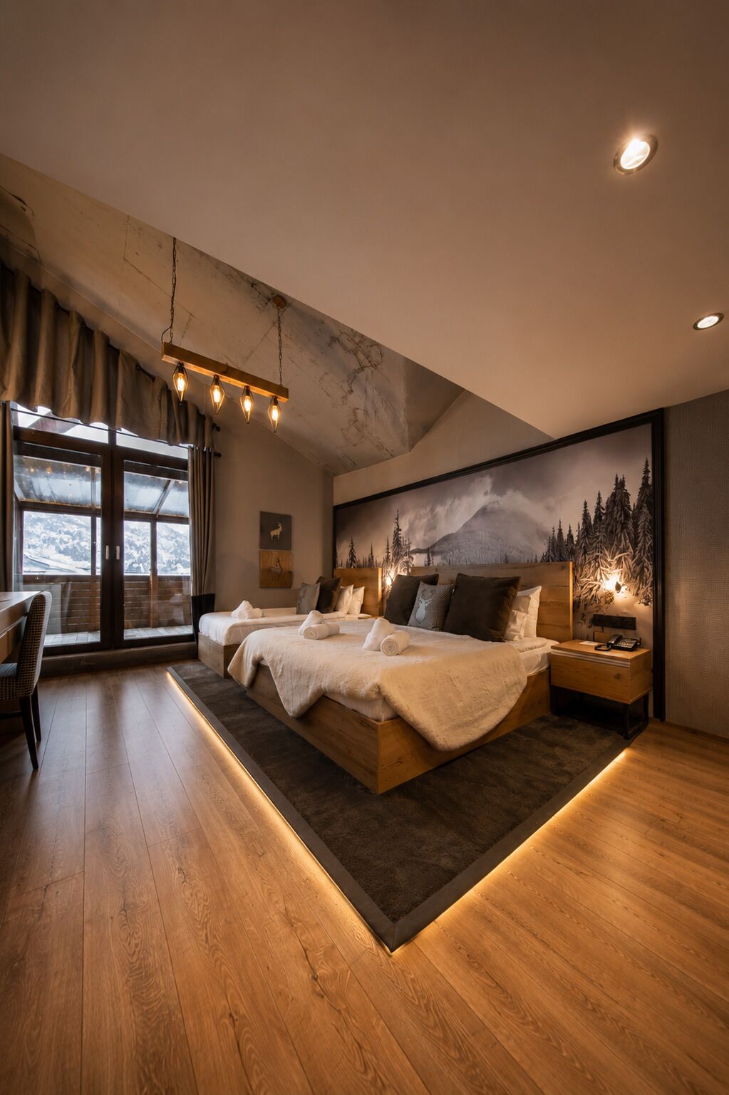 A modern bedroom with a large bed, a desk, and a view of mountains through large windows.
