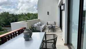 Een balkon met een witte tafel, twee rieten stoelen, een grijze bank en een grote vaas.