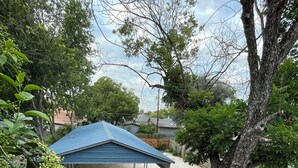 Un espacio al aire libre techado con un sendero de grava que conduce a una construcción de techo azul, rodeado de árboles y una valla de madera.