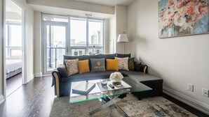 Een moderne woonkamer met een glazen salontafel, een grijze bank met kleurrijke kussens, een vloerlamp en een groot bloemschilderij aan de muur.