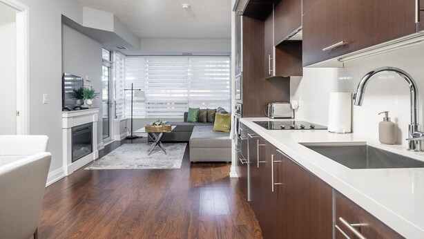 A modern kitchen with dark wood cabinets, white countertops, and stainless steel appliances. There is a dining area with a white table and chairs, and a living room with a sofa and a coffee table.