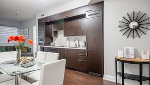 A modern kitchen with dark wood cabinets, a white countertop, and stainless steel appliances. There is a dining area with a glass table and white chairs.