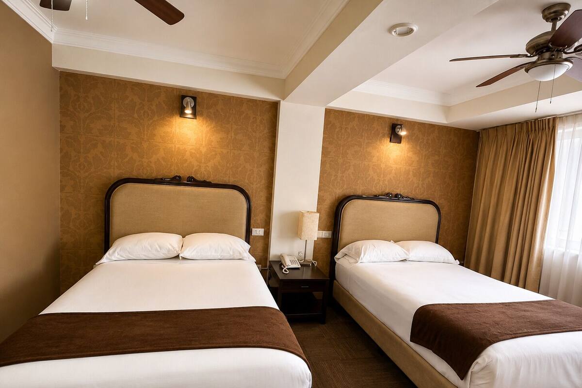 A hotel room with two beds, a ceiling fan, a nightstand with a lamp, and a window with curtains.