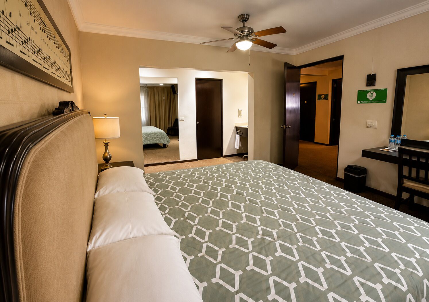 A hotel room with a bed, a desk, a chair, a ceiling fan, and a bathroom visible through an open door.