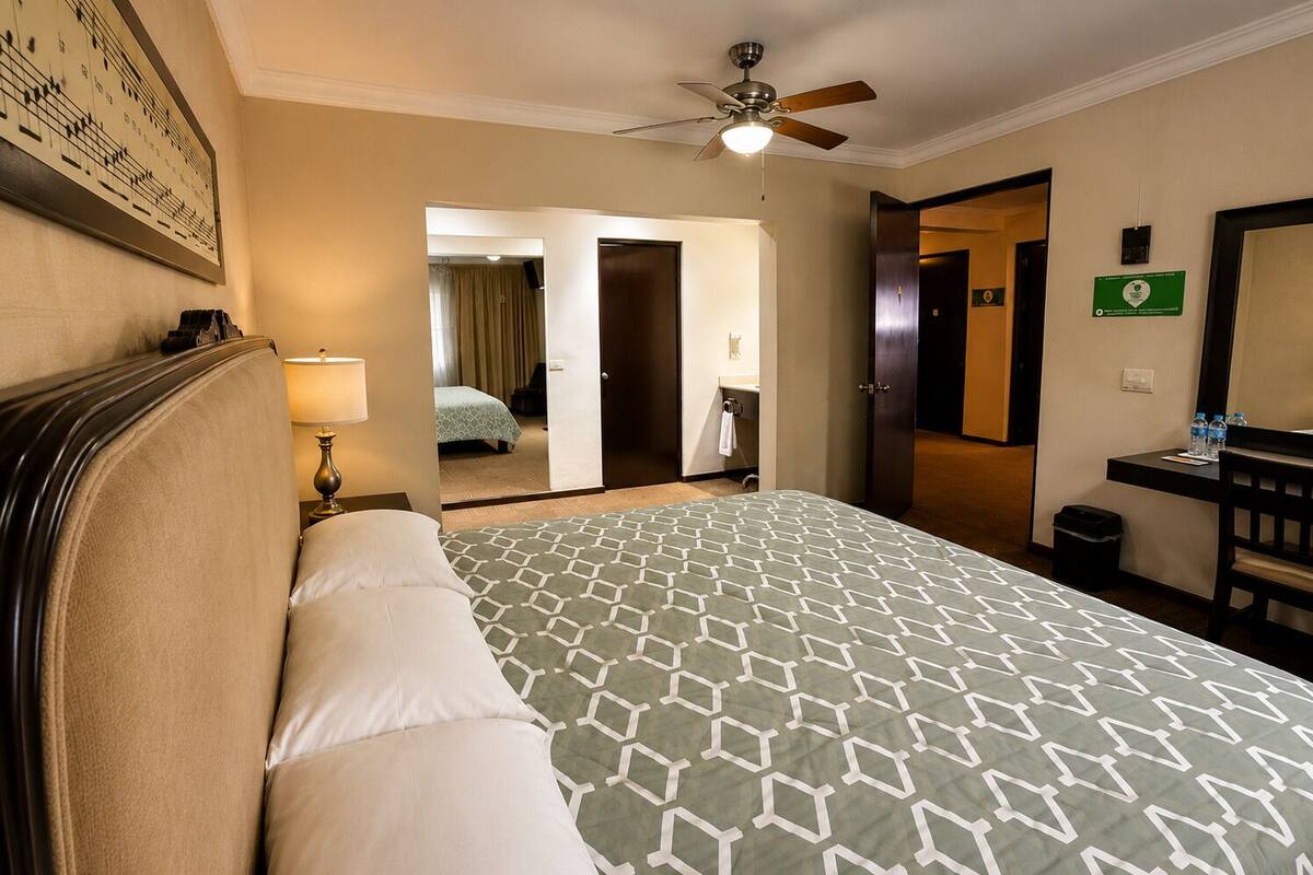 A hotel room with a bed, a desk, a chair, a ceiling fan, and a bathroom visible through an open door.