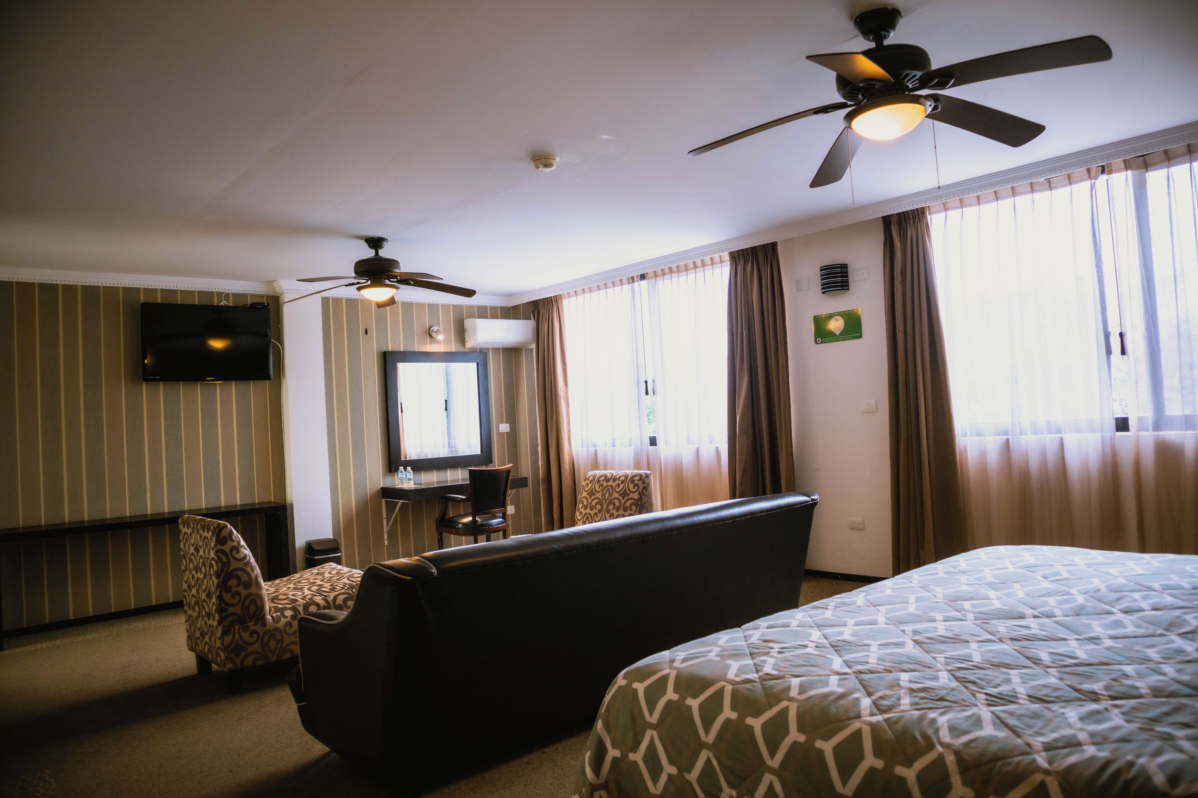 Una habitación de hotel con cama, sofá, escritorio con silla, televisor y ventilador de techo.