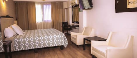 A hotel room with a bed, two chairs, a small table, a ceiling fan, and a TV mounted on the wall.