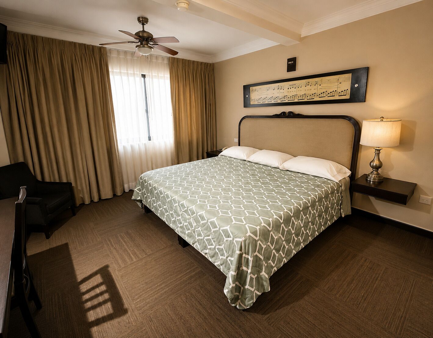A hotel room with a bed, a chair, a desk, a lamp, a ceiling fan, and a window with curtains.