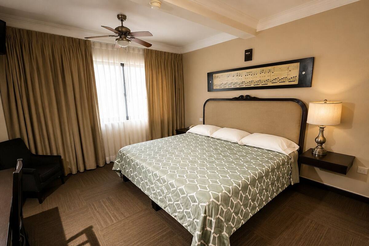 A hotel room with a bed, a chair, a desk, a lamp, a ceiling fan, and a window with curtains.