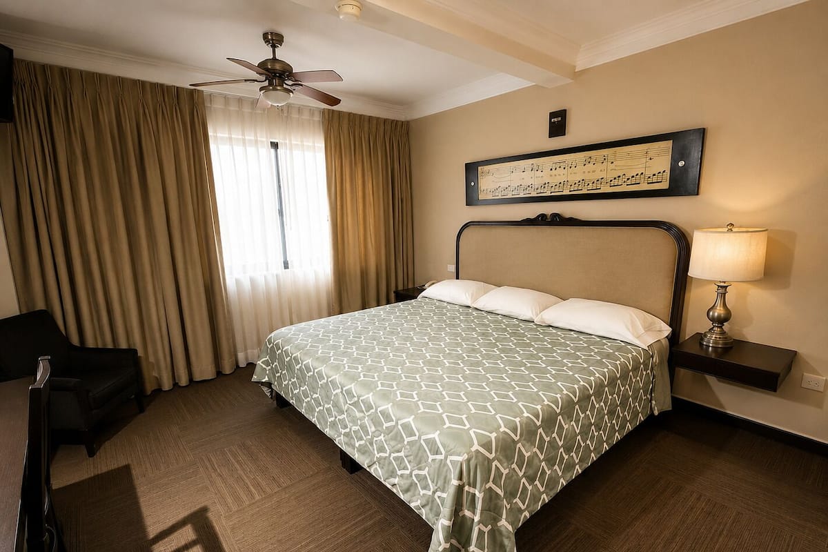 A hotel room with a bed, a chair, a desk, a lamp, a ceiling fan, and a window with curtains.