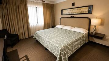 A hotel room with a bed, a chair, a desk, a lamp, a ceiling fan, and a window with curtains.