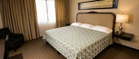 A hotel room with a bed, a chair, a desk, a lamp, a ceiling fan, and a window with curtains.