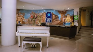 A hotel reception area with a grand piano, a large mural, and a reception desk.