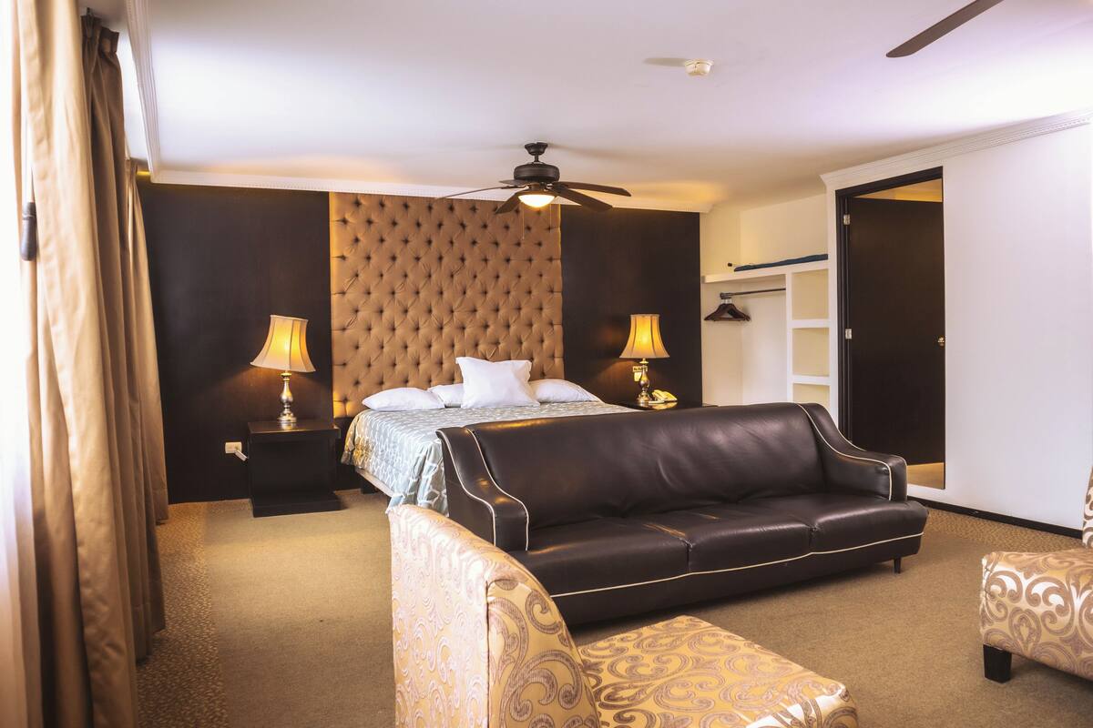 A hotel room with a bed, a sofa, a chair, a ceiling fan, and a wardrobe.
