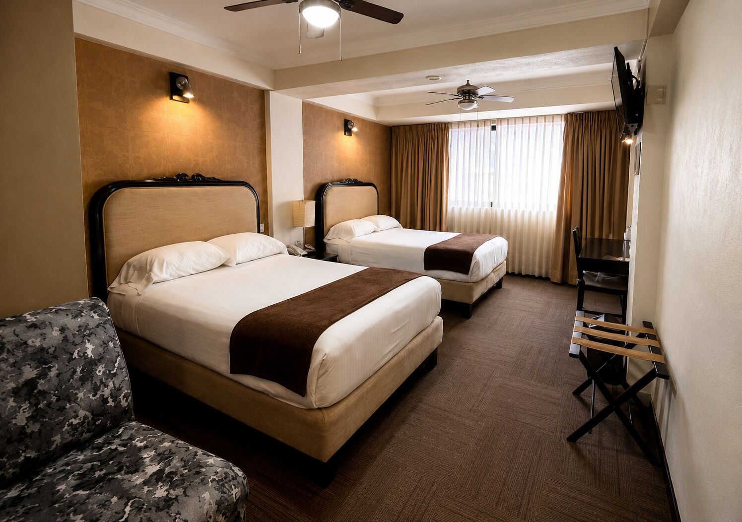 A hotel room with two beds, a desk, a chair, a ceiling fan, and a TV.