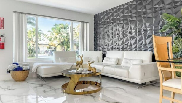 A modern living room with a white sofa, a round gold coffee table, and a geometric patterned wall. - Amazing Home w/ Heated Pool 8 mins to the Beach! (Lighthouse Point)