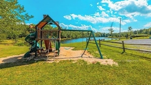 Un parque infantil con columpios y un tobogán, rodeado de césped y un lago.