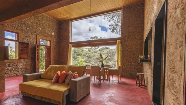 Une pièce spacieuse avec un mur en pierre, une grande fenêtre donnant sur la verdure, un canapé avec des coussins et une petite table avec des chaises.