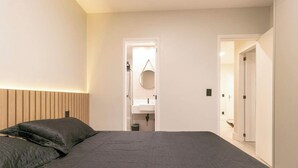 Un dormitorio moderno con cama, mesita de noche, espejo colgado y puerta hacia un baño.