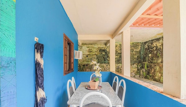A room with a blue wall, a wooden window, a table with chairs, and a view of a stone wall with greenery. - Casa em Angra dos Reis perto da Praia Jardim (Angra dos Reis)