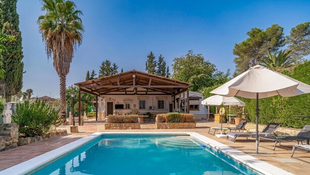 A pool area with a covered seating area, lounge chairs, and umbrellas.