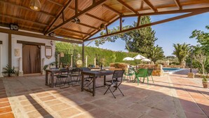 An outdoor patio with a wooden table and chairs, a pool, and a covered area with a view of trees and hills.