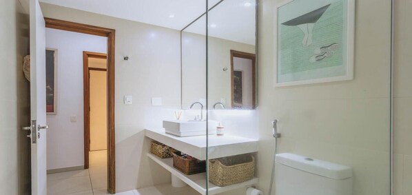 Un baño con lavamanos blanco, espejo y ducha. Hay un cuadro en la pared y una puerta que da a otra habitación.