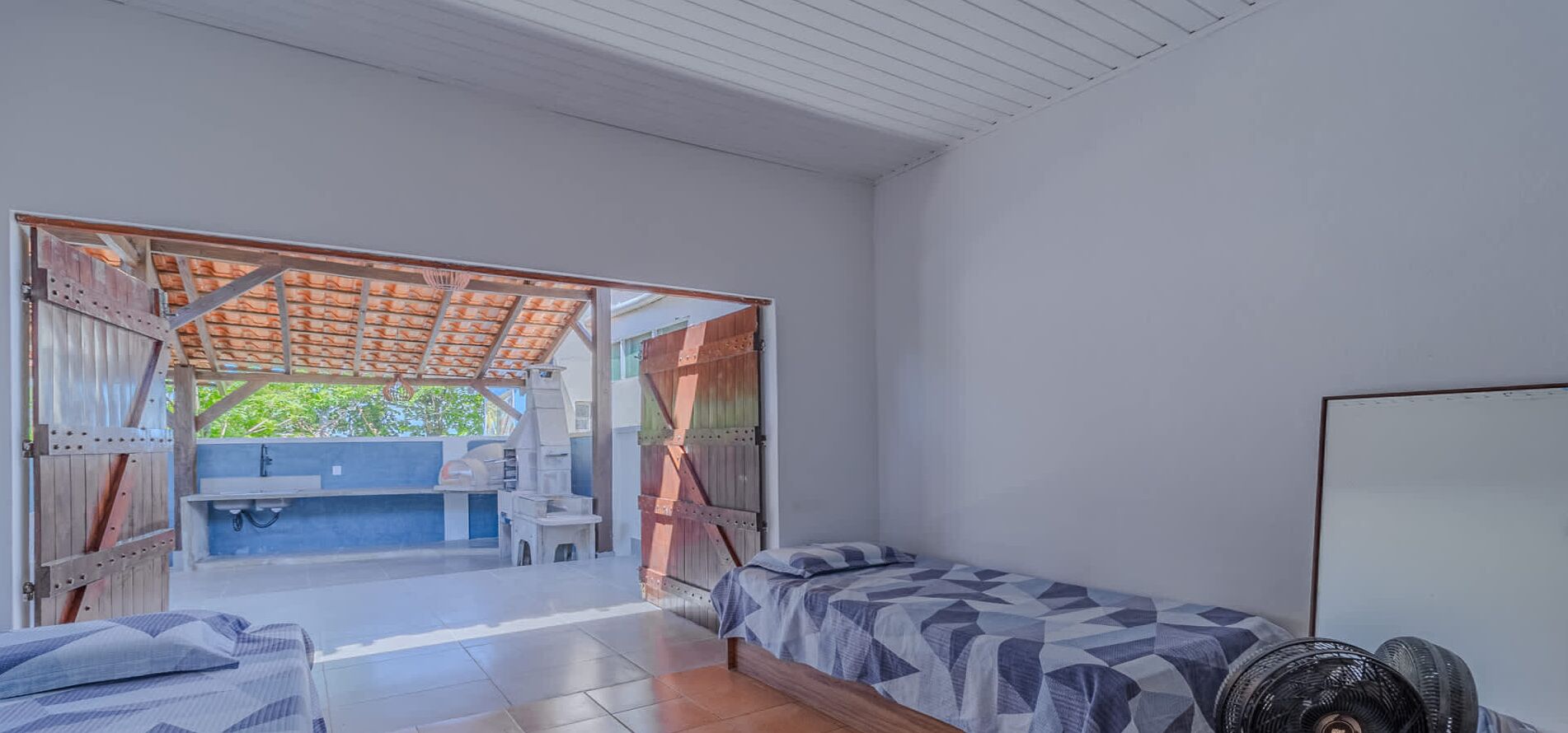 A room with a bed, a fan, and a view of an outdoor area with a wooden structure and a sink.