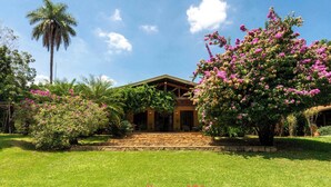 A house with a stone staircase leading to a wooden door, surrounded by lush greenery and flowering plants. - Sitio em Indaiatuba com Piscina e Churrasqueira (Campinas)