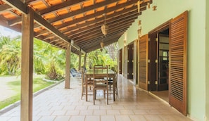 A covered patio area with a table and chairs, a view of a garden with palm trees, and a building with wooden shutters.