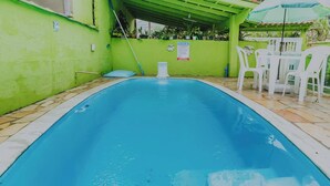 Una piscina con pared verde y un área de patio techada con sillas de plástico blanco.
