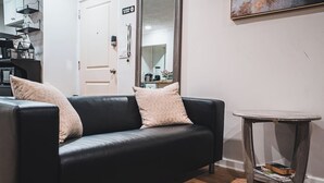 Ein schwarzes Ledersofa mit zwei Kissen, ein kleiner runder Tisch mit Zeitschriften und im Hintergrund eine Küchenzeile.