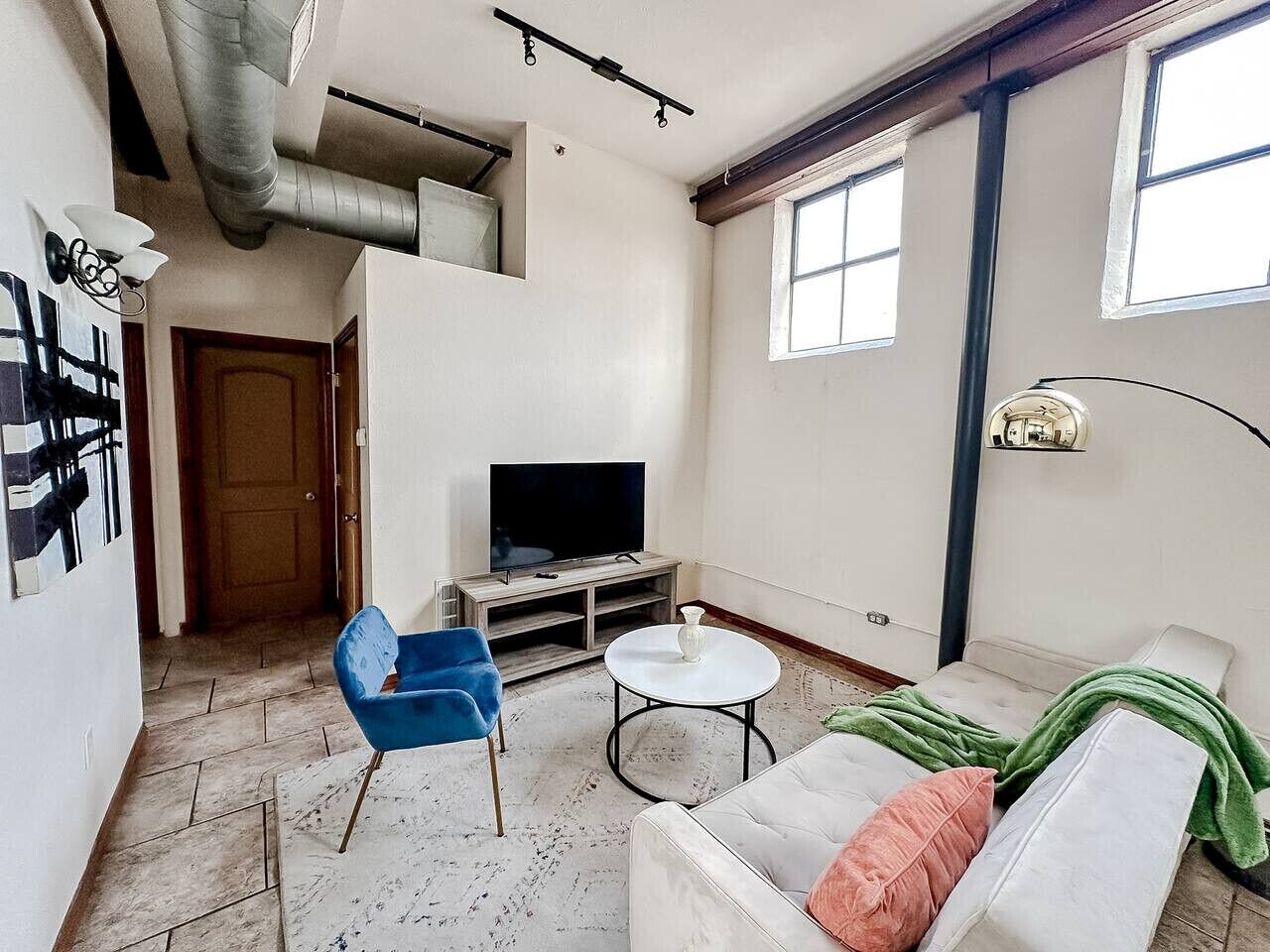 A living room with a sofa, a blue chair, a small round table, a television on a stand, and a window.