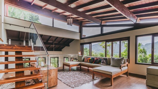 A spacious living area with a wooden staircase, a sofa with colorful cushions, and large windows offering a view of greenery and mountains. - Casa em São Sebastião com Piscina, Jacuzzi e Vista (São Sebastião)