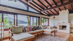 A spacious living room with a large window overlooking a scenic view, a fireplace, and a cozy seating area. - Casa em São Sebastião com Piscina, Jacuzzi e Vista (São Sebastião)