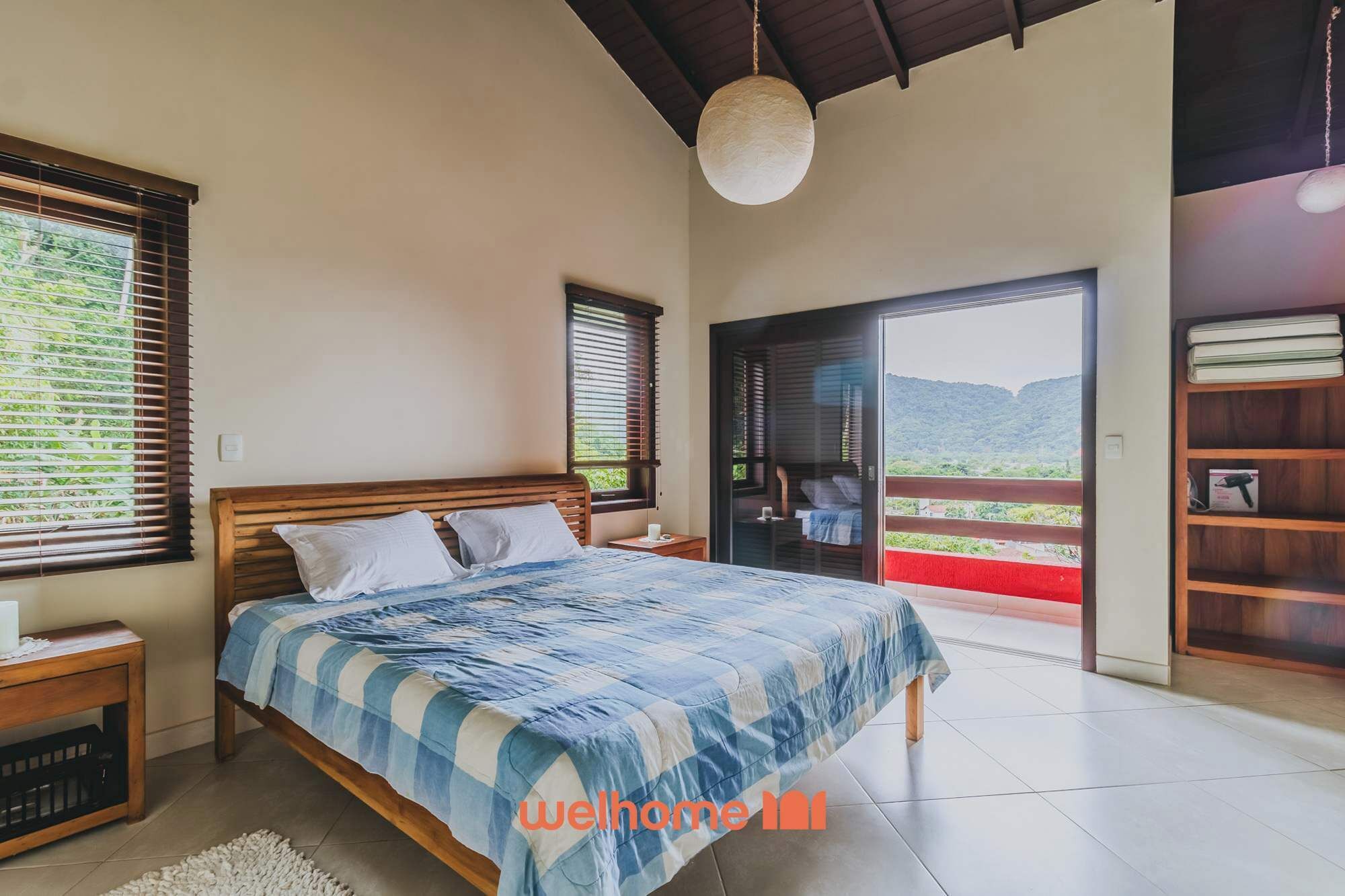 A bedroom with a bed, a nightstand, a wooden headboard, a large window with blinds, a balcony with a view of greenery, and a bookshelf.